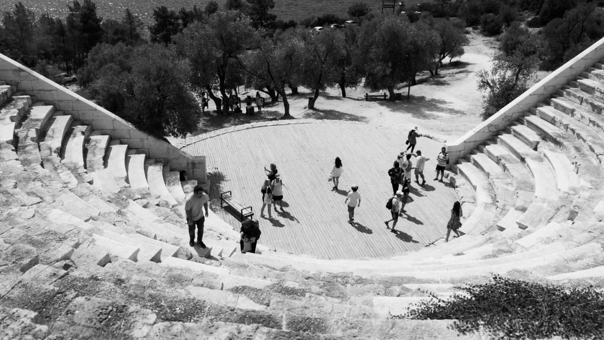 Black and white photo of people exploring an ancient Greek amphitheater by the sea.